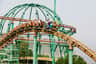 People riding a rollercoaster with green and brown tracks, set against a backdrop of trees, metal structures, and distant hills.