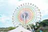A colorful ferris wheel with themed gondolas against a partly cloudy sky, with trees and a white tent in the foreground.