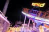 An amusement park ride named 'Disko' illuminated at night, with a carousel and skyscraper in the background.