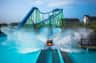 A log flume ride splashing into water with a vibrant roller coaster in the background under a clear blue sky.