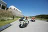 Children riding downhill on luge carts on a paved track under a clear blue sky, with a building and greenery in the background.