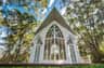 A small white chapel with arched windows surrounded by tall trees under a blue sky with scattered clouds.