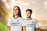A woman and a man smiling, wearing Real Madrid jerseys, in a brightly lit stadium with a sunset sky. Real Madrid World logo in the corner.