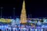 A brightly lit outdoor ice skating rink surrounded by festive holiday lights and decorations, including a large illuminated tree.